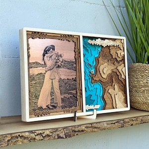 May include: A wooden framed photo and topographic map display. The left side features a sepia-toned photo of a couple embracing. The right side has a layered blue and brown topographic map with a heart and the text "April 13, 2017".