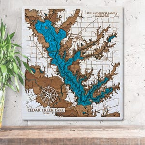 May include: A wooden wall art piece depicting a detailed map of Cedar Creek Lake in Texas. The lake is rendered in shades of blue, with surrounding land in brown and white. Includes a compass rose and text: "THE ANDJELICH FAMILY" and "Established 2020".
