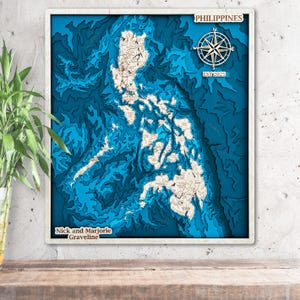 May include: A framed, layered wooden map of the Philippines, showcasing the islands in shades of blue and beige. The map includes a compass rose and the text "PHILIPPINES" and "EST 2025". The names "Nick and Marjorie Graveline" are also displayed.