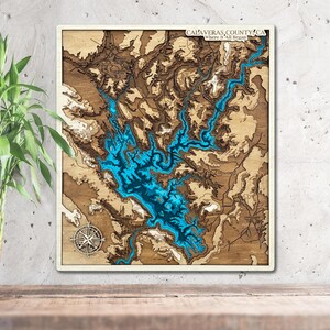 May include: A wooden map of Calaveras County, California, with a blue lake in the center. The map is framed in a light wood frame and has the text "Calaveras County, CA Where It All Began" at the top.