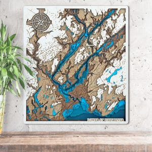 May include: A layered wooden map of the Lower St. John River, featuring a compass rose in the upper left corner. The map showcases the river's course in blue, with surrounding land in shades of brown and white. The text "LOWER ST. JOHN RIVER" is at the bottom.