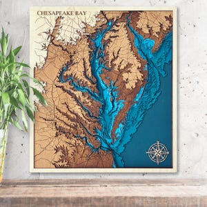 May include: A wooden, layered map of Chesapeake Bay, featuring detailed topography in shades of brown and blue. The text "CHESAPEAKE BAY" is at the top. A compass rose is in the lower right corner. The map is framed in a light-colored wood.