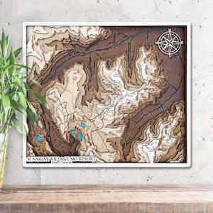 May include: A layered wooden map of Sunshine Village Ski Resort in Banff, Canada. The map features detailed topographical contours in shades of brown and beige, with blue accents for lakes and rivers. A compass rose is in the upper right corner.
