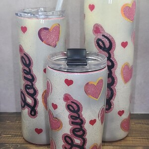 Puede incluir: Tres vasos blancos con acabado brillante y corazones rosas con contornos dorados. Los vasos tienen la palabra "Love" escrita en ellos en cursiva negra.