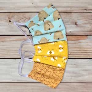 May include: Three reusable face masks with bee-themed patterns. The top mask is light blue with brown beehives and yellow bees. The middle mask is yellow with white bees and brown beehives. The bottom mask is yellow with a honeycomb pattern.