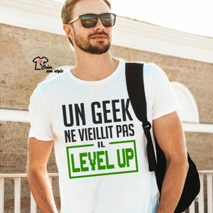 Peut inclure: T-shirt blanc avec un texte noir qui dit "Un Geek Ne Vieillit Pas Il Level Up" avec le mot "Level Up" en vert.