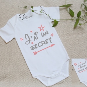 Op de afbeelding: Witte babybody met de tekst "J'AI UN SECRET" in grijs, met roze sterren en een pijl. Een kleinere body toont de tekst "JE SERAI BIENTÔT GRANDE SOEUR". Een schattig babykledingstuk.