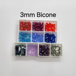 K&ouml;nnte beinhalten: Ein wei&szlig;es Kunststoff-Sortierfach mit neun F&auml;chern, gef&uuml;llt mit 3 mm Bicone-Perlen in verschiedenen Farben wie Rot, Magenta, Orange, Hellblau, Mittelblau, Dunkelblau, Schwarz, Lavendel, Violett und Grau mit dem Text 3mm Bicone.
