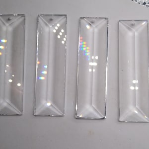 May include: Four clear glass prism pendants, each with a rectangular shape and a beveled edge. The prisms are arranged in a row on a white surface.
