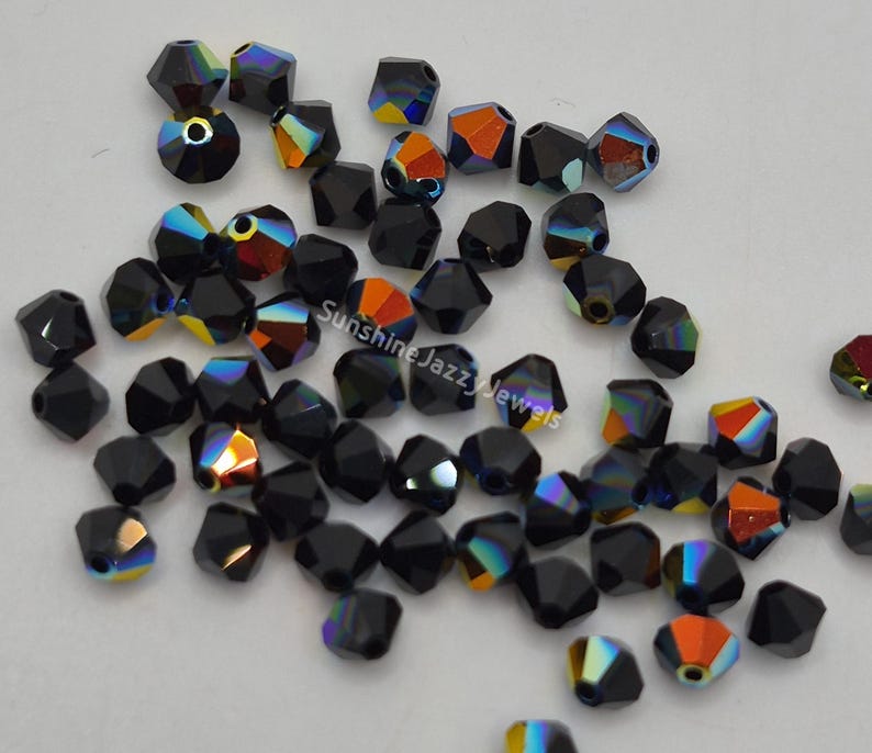 May include: A collection of faceted black beads with iridescent color flashes of orange, yellow, and blue. The beads are scattered on a white surface. The beads are likely for jewelry making.
