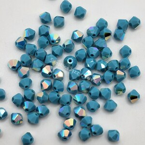 May include: A collection of faceted, teal-colored beads with iridescent flashes. The beads are scattered across a white surface, showcasing their geometric shapes and light-catching qualities. The beads are ideal for jewelry making.