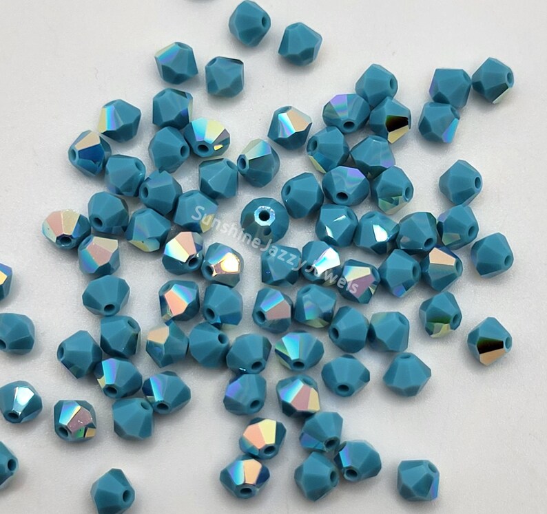May include: A collection of faceted, teal-colored beads with iridescent flashes. The beads are scattered across a white surface, showcasing their geometric shapes and light-catching qualities. The beads are ideal for jewelry making.