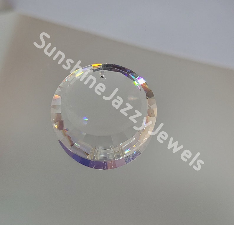 May include: A clear crystal pendant with a faceted design and a hole at the top for hanging. The pendant is round and has a rainbow iridescent sheen.