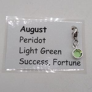 May include: A silver charm with a light green peridot stone. The charm is labeled "August, Peridot, Light Green, Success, Fortune".