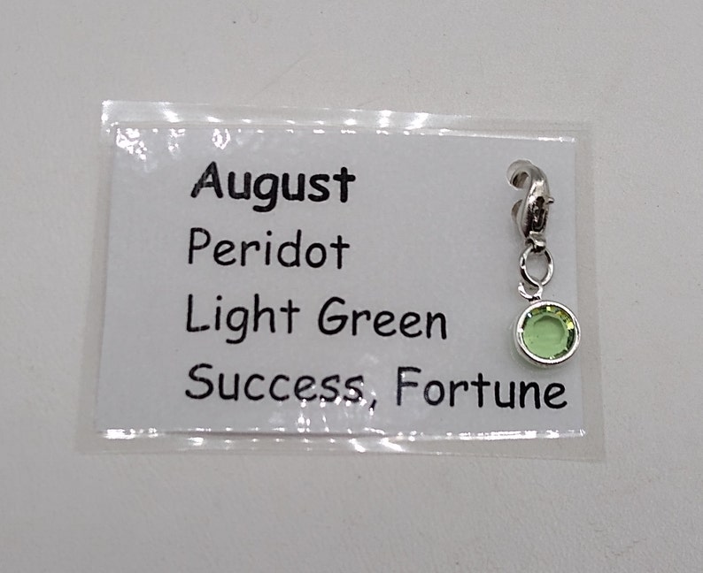 May include: A silver charm with a light green peridot stone. The charm is labeled "August, Peridot, Light Green, Success, Fortune".