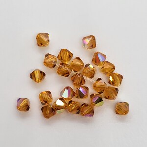 May include: A collection of square-cut, amber-colored crystal beads. Each bead has multiple facets that reflect light, creating a shimmering effect. The beads are scattered on a white surface, showcasing their color and shape.