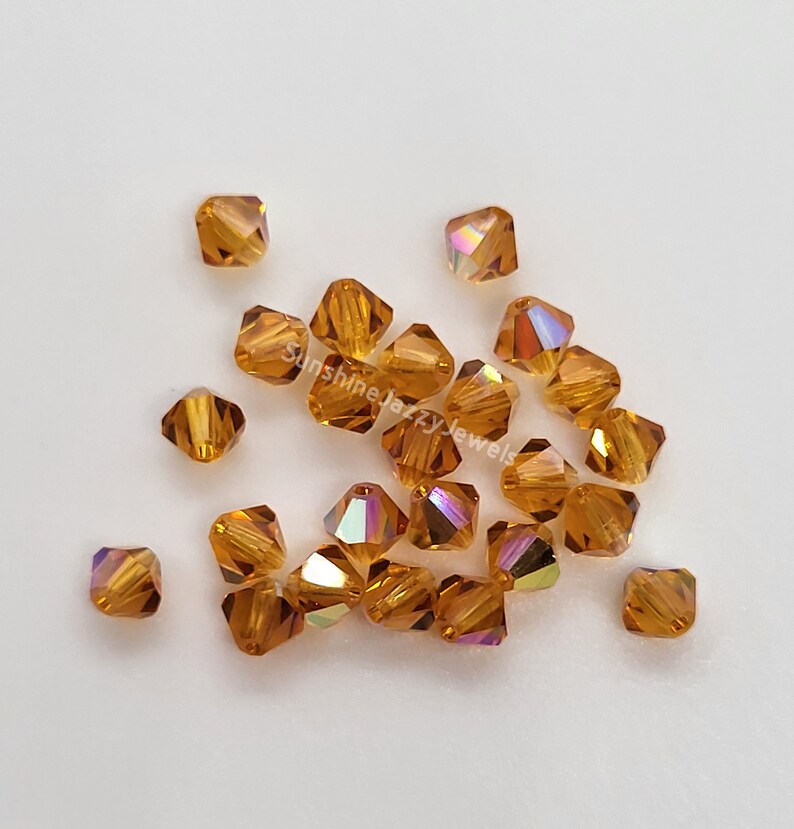 May include: A collection of square-cut, amber-colored crystal beads. Each bead has multiple facets that reflect light, creating a shimmering effect. The beads are scattered on a white surface, showcasing their color and shape.