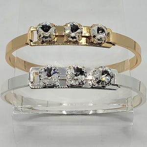 May include: Two bangle bracelets, one gold and one silver, each with three clear crystal stones set in a metal bar.