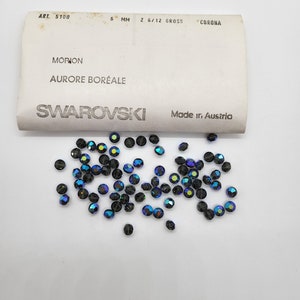 May include: A close-up of a pile of small, iridescent blue and black glass beads. The beads are labeled "Swarovski" and "Morion Aurore Boreale".