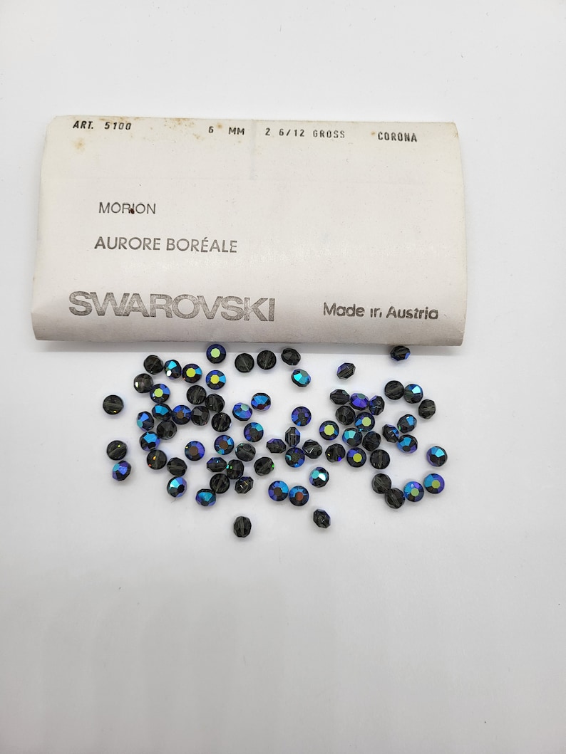 May include: A close-up of a pile of small, iridescent blue and black glass beads. The beads are labeled "Swarovski" and "Morion Aurore Boreale".