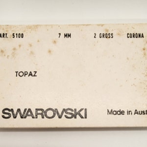 Puede incluir: Una caja blanca con texto negro que dice "ART. 5100 7 MM 2 GROSS CORONA TOPAZ SWAROVSKI Made in Austria".