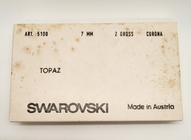 Puede incluir: Una caja blanca con texto negro que dice "ART. 5100 7 MM 2 GROSS CORONA TOPAZ SWAROVSKI Made in Austria".