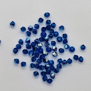 K&ouml;nnte beinhalten: Eine Ansammlung kleiner, facettierter, tiefblauer Kristallperlen, die auf einer glatten Oberfl&auml;che verstreut sind. Jede Perle hat einen mehrseitigen geometrischen Schliff, der das Licht reflektiert und einen funkelnden Effekt erzeugt. Der Text SunshineJazzyJewels ist sichtbar.