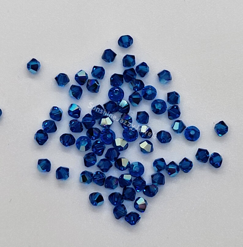 K&ouml;nnte beinhalten: Eine Ansammlung kleiner, facettierter, tiefblauer Kristallperlen, die auf einer glatten Oberfl&auml;che verstreut sind. Jede Perle hat einen mehrseitigen geometrischen Schliff, der das Licht reflektiert und einen funkelnden Effekt erzeugt. Der Text SunshineJazzyJewels ist sichtbar.