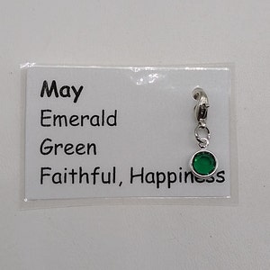 May include: A silver charm with a green emerald stone. The charm has a clip on the top and says "May, Emerald, Green, Faithful, Happiness" on a white card.