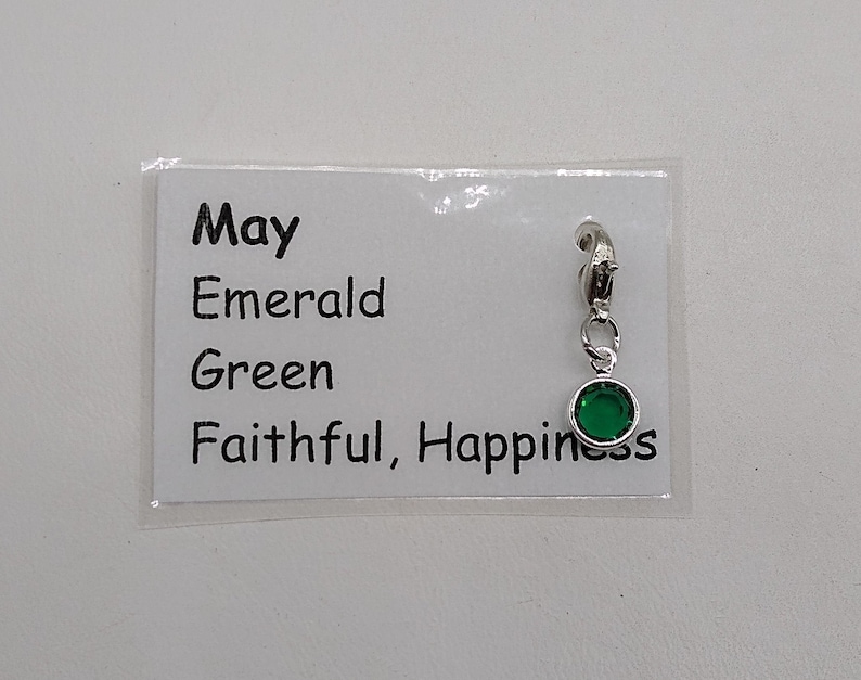 May include: A silver charm with a green emerald stone. The charm has a clip on the top and says "May, Emerald, Green, Faithful, Happiness" on a white card.