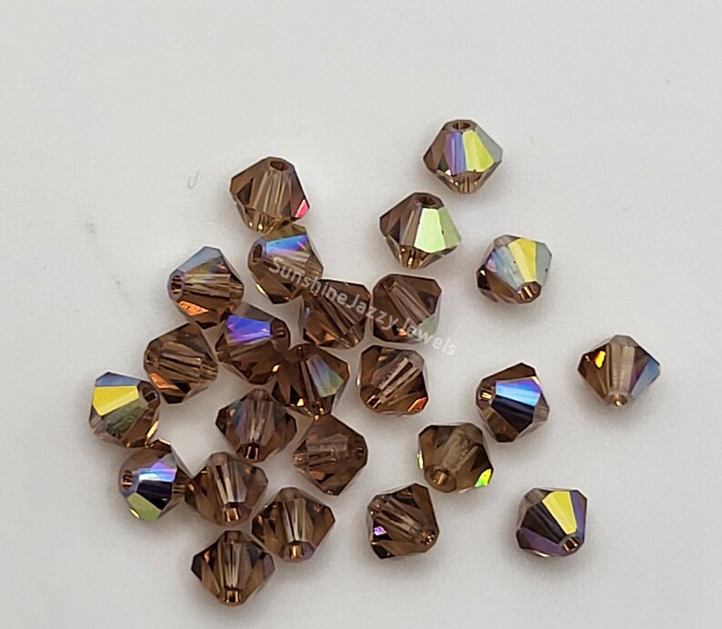 May include: A collection of faceted, brown crystal beads with iridescent color shifts. The beads are scattered on a white surface. The text "Sunshine Jazzy Jewels" is visible in the image.