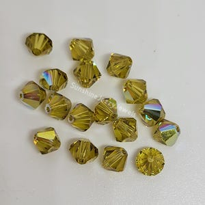May include: A collection of faceted, golden-yellow crystal beads. The beads have a square shape with multiple facets that catch the light, creating a shimmering effect. Some beads display iridescent colors. The text "Sunshine Jazzy Jewels" is visible.