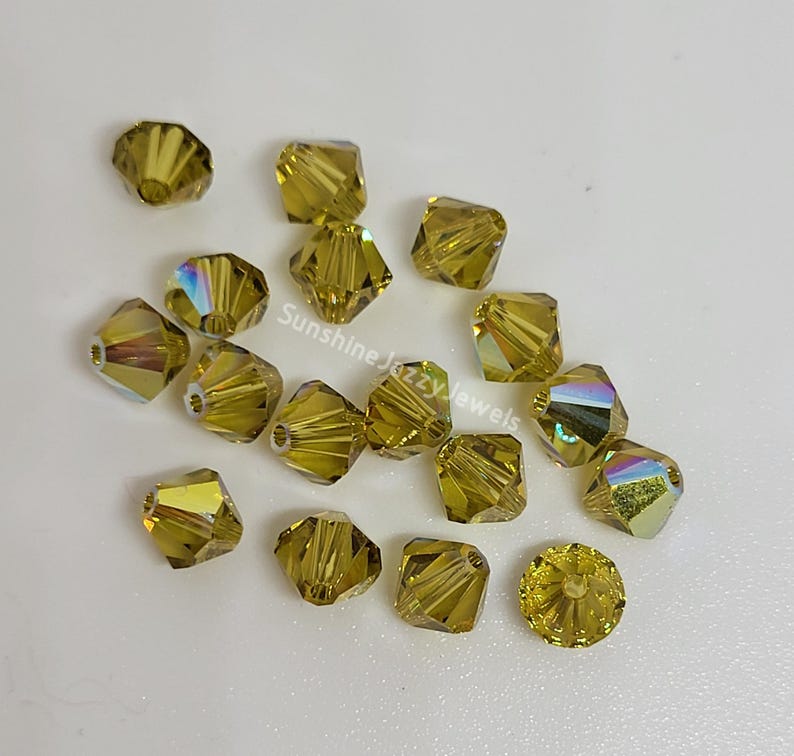 May include: A collection of faceted, golden-yellow crystal beads. The beads have a square shape with multiple facets that catch the light, creating a shimmering effect. Some beads display iridescent colors. The text "Sunshine Jazzy Jewels" is visible.