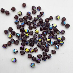 May include: A collection of faceted, dark purple beads with iridescent, multi-colored reflections. The beads are scattered across a white surface, showcasing their geometric shapes and light-catching qualities. Ideal for jewelry making.