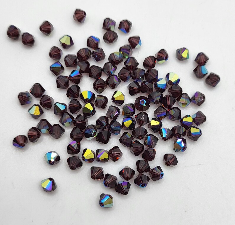 May include: A collection of faceted, dark purple beads with iridescent, multi-colored reflections. The beads are scattered across a white surface, showcasing their geometric shapes and light-catching qualities. Ideal for jewelry making.