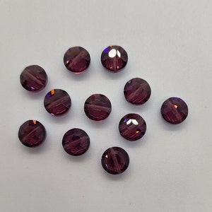 May include: Eleven round, faceted beads in a deep purple colour. The beads have a sparkling, crystal-like appearance, reflecting light with multiple facets. They are scattered on a white surface, showcasing their colour and shape.
