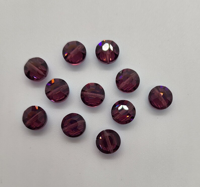May include: Eleven round, faceted beads in a deep purple colour. The beads have a sparkling, crystal-like appearance, reflecting light with multiple facets. They are scattered on a white surface, showcasing their colour and shape.