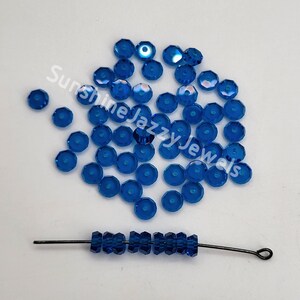 May include: A large quantity of small, round, blue glass beads with a hole in the center. The beads are scattered on a white surface. A single strand of the same beads is strung on a thin wire.