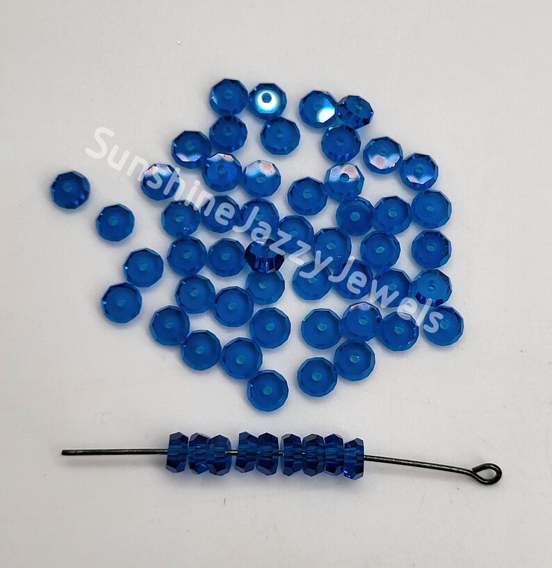 May include: A large quantity of small, round, blue glass beads with a hole in the center. The beads are scattered on a white surface. A single strand of the same beads is strung on a thin wire.