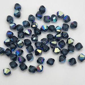 May include: A collection of faceted, dark blue beads with iridescent highlights. The beads are scattered across a white surface, showcasing their multiple facets and color variations. These beads are suitable for jewelry making and crafting.