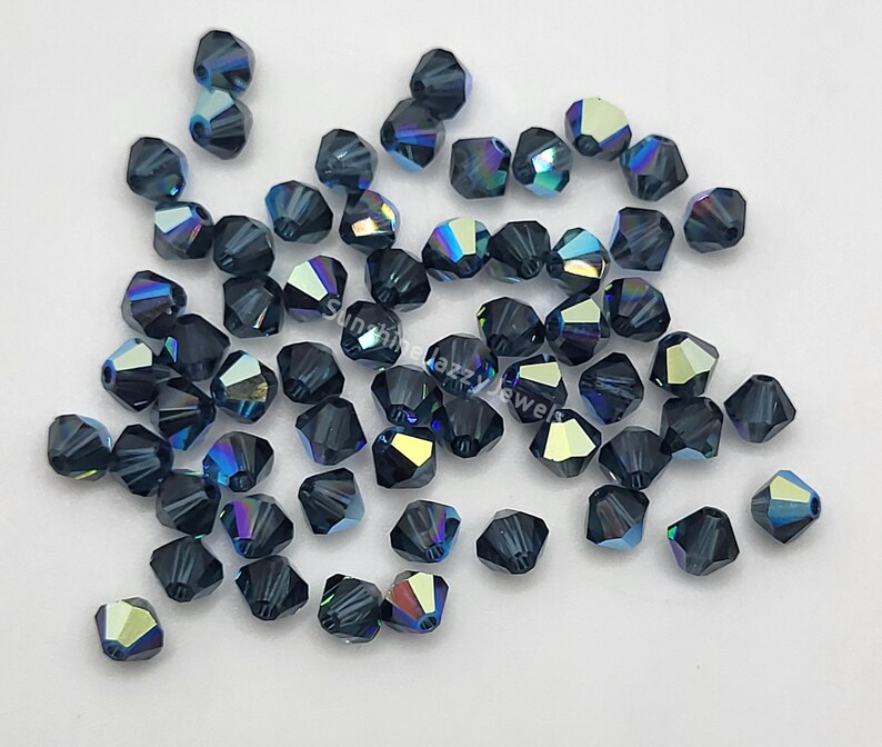 May include: A collection of faceted, dark blue beads with iridescent highlights. The beads are scattered across a white surface, showcasing their multiple facets and color variations. These beads are suitable for jewelry making and crafting.