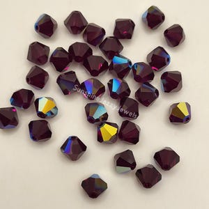 May include: A collection of faceted, dark purple beads with iridescent color shifts. The beads are scattered, showcasing their multiple sides and the play of light. The text "Sunshine Jazzy Jewels" is visible on the image.