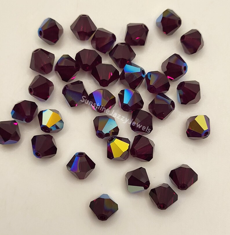 May include: A collection of faceted, dark purple beads with iridescent color shifts. The beads are scattered, showcasing their multiple sides and the play of light. The text "Sunshine Jazzy Jewels" is visible on the image.