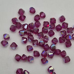 May include: A scattering of faceted, deep magenta beads with iridescent flashes. The beads are scattered across a light surface. The text "Sunshine Jazzy Jewels" is visible on the surface.