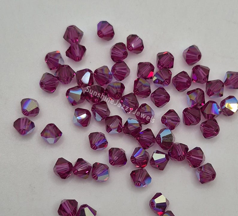 May include: A scattering of faceted, deep magenta beads with iridescent flashes. The beads are scattered across a light surface. The text "Sunshine Jazzy Jewels" is visible on the surface.