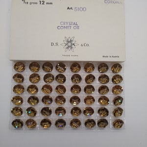 May include: A clear plastic container filled with 144 gold-toned crystal beads. The container has a white label with the text "CORONA Art. 5100 CRYSTAL COMET OR D.S. & Co. TRADE MARK Made in Austria".