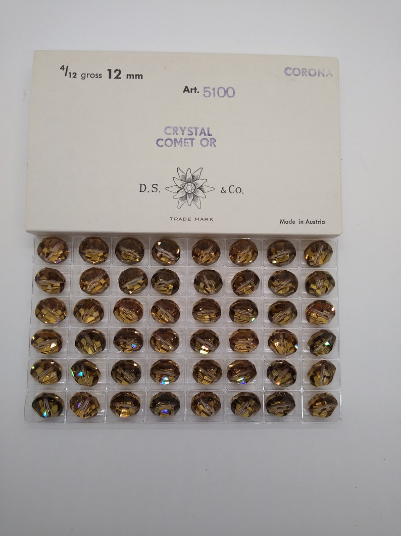 May include: A clear plastic container filled with 144 gold-toned crystal beads. The container has a white label with the text "CORONA Art. 5100 CRYSTAL COMET OR D.S. & Co. TRADE MARK Made in Austria".