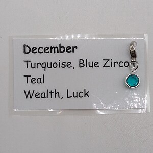 May include: A silver charm with a turquoise blue gemstone. The charm is labeled "December, Turquoise, Blue Zircon, Teal, Wealth, Luck".
