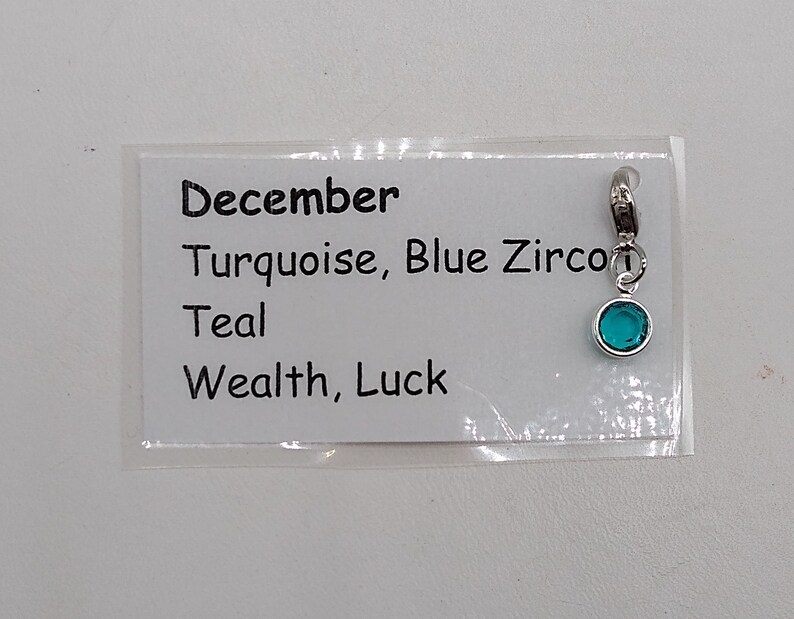 May include: A silver charm with a turquoise blue gemstone. The charm is labeled "December, Turquoise, Blue Zircon, Teal, Wealth, Luck".