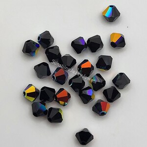 May include: A collection of black, faceted beads with iridescent color flashes of orange, yellow, and purple. The beads are scattered on a white surface. The text "Sunshine Jazzy Jewels" is visible on the image.
